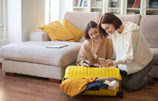 Happy european young women with smartphone, tickets and suitcase with clothes, preparing for travel together in living room interior. Relationships, friendship at home, travel together and vacation photo