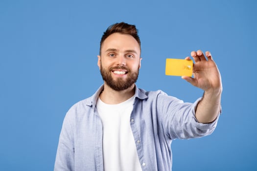 Positive millennial guy showing credit card and smiling on blue studio background. Young bearded man promoting contactless payment, using electronic money during coronavirus outbreak photo