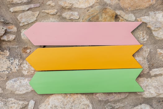 Vibrant wooden mockup arrows in pink, yellow, and green are displayed against a textured stone wall outdoors under natural light. photo