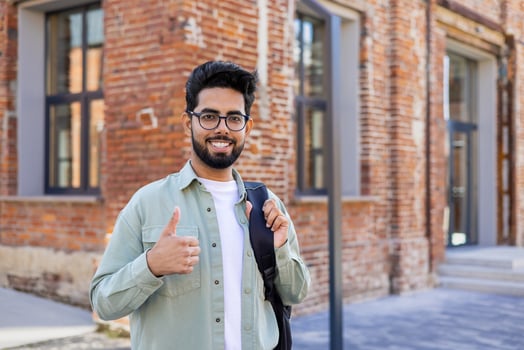 Portrait of a young Indian male student, freelancer standing outside on the street with a backpack, smiling at the camera and showing the super sign with his finger. photo