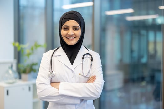 A smiling female doctor wearing a hijab and medical coat stands with arms crossed in a modern office setting. photo