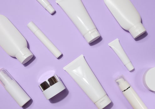 A variety of white plastic tubes and bottles with cosmetics arranged randomly on a lavender background. Top view photo