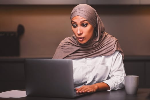 Shocked Muslim Woman Looking At Laptop Computer Browsing Internet And Reading Shocking News Online Sitting In Modern Kitchen At Home, Wearing Hijab. Omg Concept photo