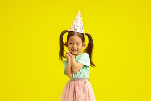 Asian little girl in party hat smiling coyly, cute placed palms of hands in front of herself face, looks like as if begging for a desired gift or makes a wish on herself birthday, stands over yellow photo
