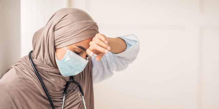 Stressed muslim woman doctor in protective face mask touching her sweating forehead, feeling exhausted after difficult working day, sitting by wall at clinic, copy space. Doctors and COVID-19 pandemic photo