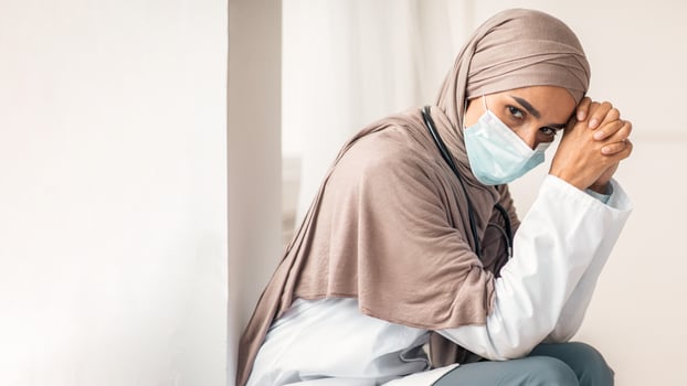 Pandemic outbreak at hospitals and clinics. Pensive muslim woman doctor sitting next to wall in her cabinet at clinic, having short break during working shift, wearing protective face mask, free space photo