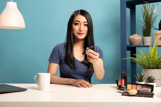Asian beauty influencer seated at desk, filming a makeup tutorial. Female vlogger holds a lipstick, presenting it to camera and recording a to engage her subscribers with expert cosmetic advice. photo