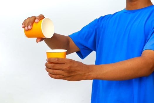 Person in blue shirt pours liquid from one paper cup to another against white background photo