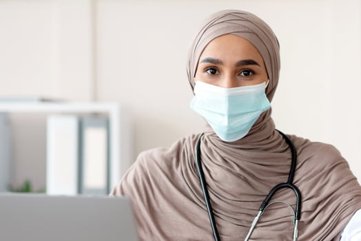 Muslim woman doctor in protective face mask and hijab sitting at workdesk with modern laptop in her cabinet in clinic or hospital, taking anamnesis, having appointment during pandemic, copy space photo
