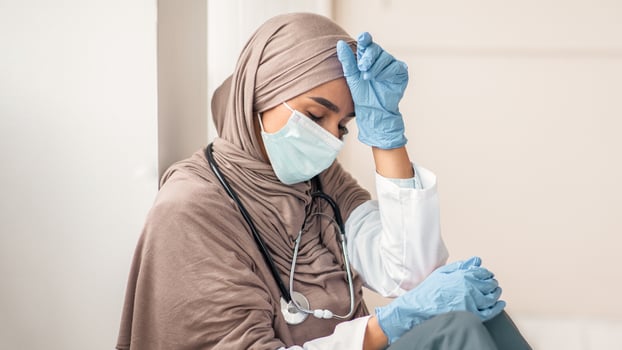 Upset woman in hijab doctor having difficult day at clinic, wearing protective gloves and face mask, sitting next to wall in her office and resting, empty space. Sad female doctor feeling exhausted photo
