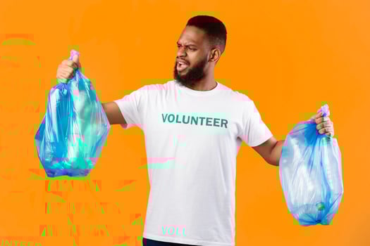 Displeased African American Volunteer Guy Holding Two Plastic Bags Full Of Garbage Litter And Bottles Posing In Studio Over Yellow Background. Ecology Problems And Pollution, Sort Your Waste Concept photo