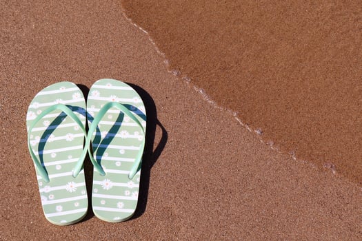 A pair of light green flip-flops with a daisy pattern rests on the wet sandy beach as a gentle wave approaches. The photo captures the essence of a relaxing summer day.
