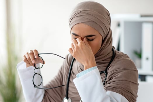 Muslim doctor young woman in hijab with stethoscope having eyesight problem while working with laptop at clinic, holding glasses and rubbing her nose bridge with closed eyes, copy space photo