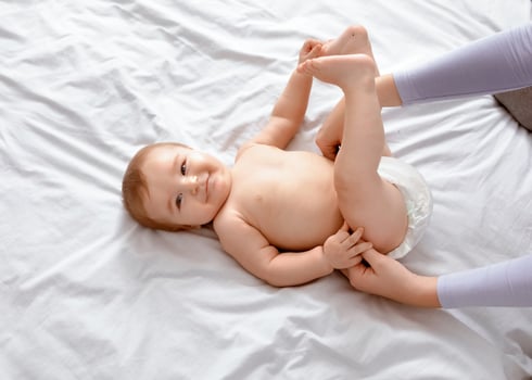 Unrecognizable mother changing her smiling little baby's diaper on white, top view. Cute toddler blonde boy having fun while his mom dressing him up, playing with his feet, closeup, empty space. photo