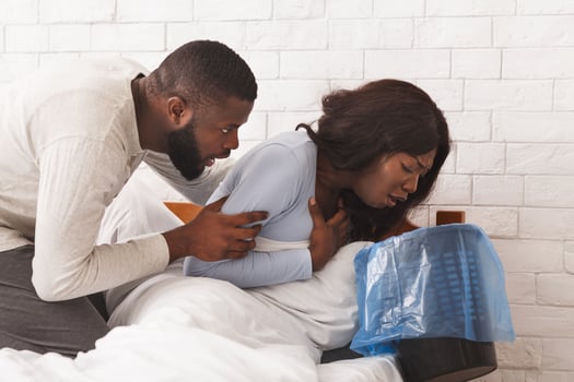 Morning sickness. Young pregnant african woman sitting on bed, holding trash, feeling nauseous during pregnancy, her man comforting her photo