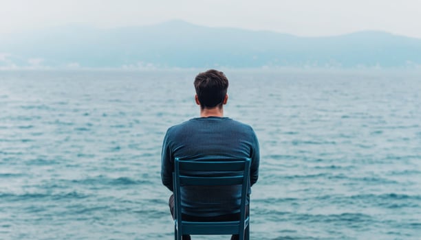 Lonely adult man sitting alone by the sea at sunset looking away, back view. photo