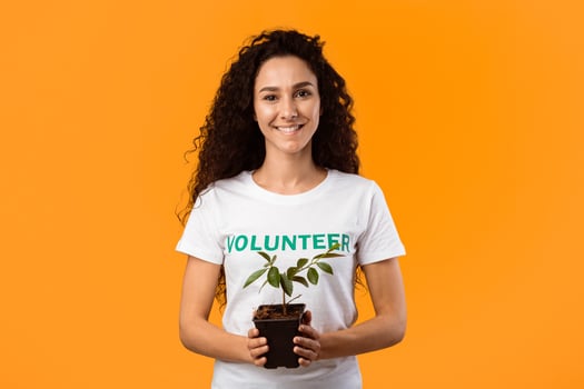 Female Volunteer Holding Pot With Green Plant Smiling To Camera Standing On Yellow Background. Protection Of Environment And Nature, Ecology Concept. Studio Shot photo