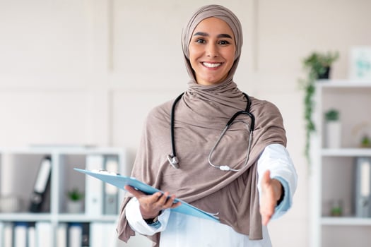Happy female doctor young woman in hijab with medical folder greeting someone in her office, copy space. Muslim woman therapist holding medical chart and giving hand, clinic interior photo