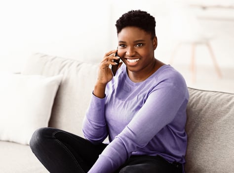 Happy cheerful young black woman talking on the phone at home, smiling lady making call by cellphone sitting on sofa at home, having pleasant or funny conversation speaking by mobile photo