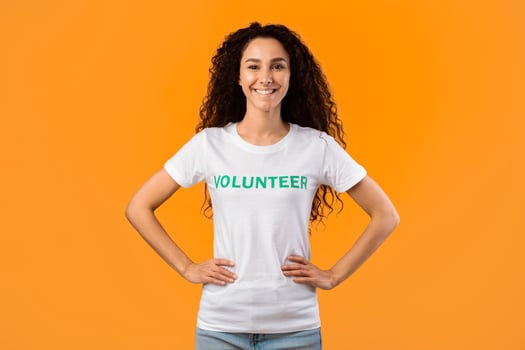 Enthusiastic Volunteer Woman Posing With Hands On Her Hips Smiling To Camera Standing Ready For Voluntary Work Over Yellow Studio Background. Female Activist Portrait. Volunteering Occupation Concept photo