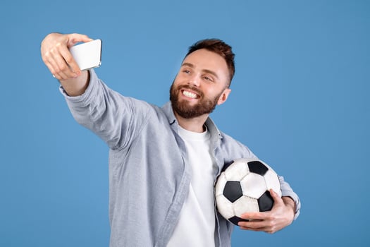 Positive millennial guy with soccer ball taking selfie on smartphone over blue studio background. Joyful football fan making photo of himself before championship, rooting for his favorite team
