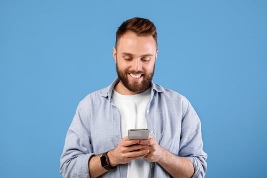 Online life soncept. Funky bearded man using cellphone on blue studio background. Positive young guy with modern gadget surfing internet, watching videos, posting in social media photo