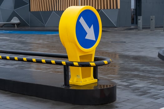 A bright yellow directional sign with an arrow points left at a contemporary city intersection. The wet pavement reflects the surroundings, highlighting the urban design and rainy weather photo