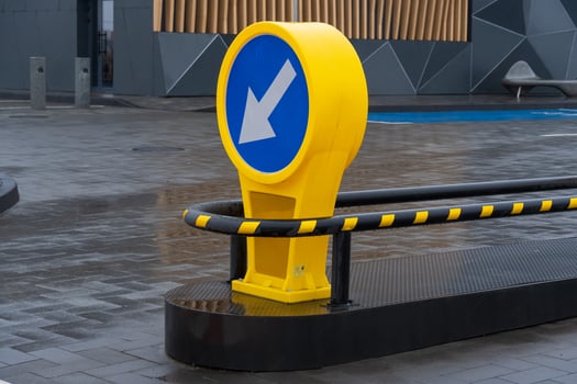 A bold yellow directional sign with a blue circular background indicating leftward movement stands on a sleek pavement in an urban setting. The surroundings showcase contemporary architecture photo