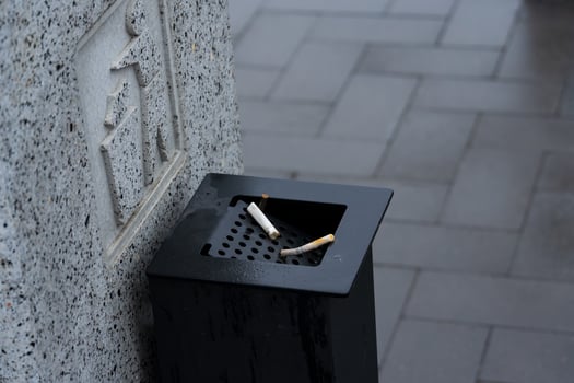 A black waste container filled with discarded cigarette butts stands on a wet pavement under gray skies in a bustling city photo