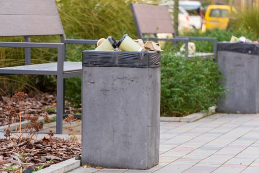 In a vibrant city park during a sunny afternoon, a concrete rubbish container overflows with trash bags photo