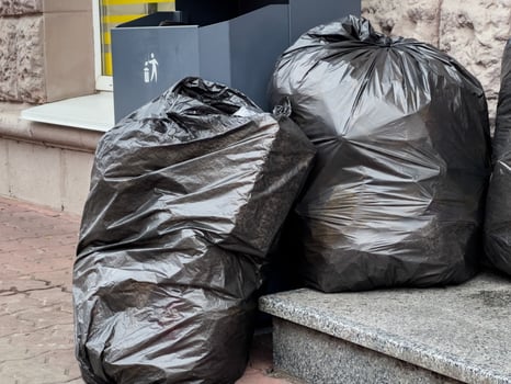 Black garbage bags lean against a clean, modern trash container on a city sidewalk. The sunlight highlights the need for environmental awareness and responsible waste management in urban areas photo