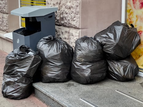 Several large black trash bags piled up beside a recycling bin on a city sidewalk. The setting emphasizes urban waste management and environmental conservation efforts in action photo