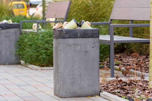 A concrete trash bin stands filled with waste bags, surrounded by greenery and benches in a bustling urban park. This spot reflects both convenience and environmental responsibility photo