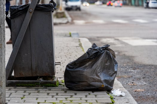 A garbage container stands near the curb of an urban street. Black trash bags are placed next to the bin, highlighting the issue of waste management and recycling photo