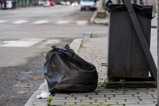A black garbage bag sits on the curb beside a recycling bin, illustrating daily urban waste management efforts. The setting captures a typical city street with vehicles passing by photo