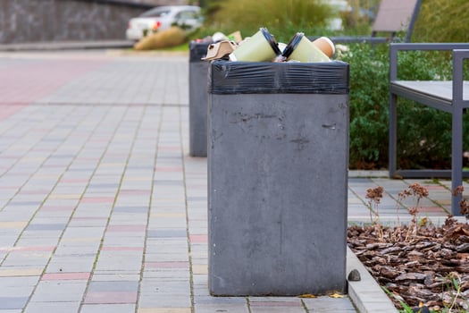 A concrete garbage container is filled with trash bags on a busy city sidewalk. Nearby plants and benches are visible, highlighting urban waste management challenges photo