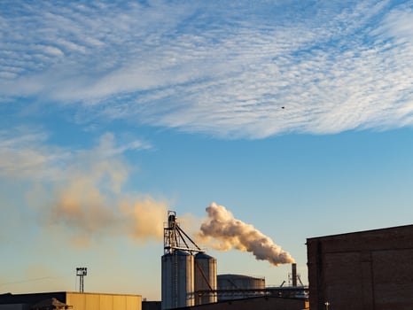 Billowing smoke emerges from smokestacks at an industrial facility during sunset, casting a cloudy backdrop over the manufacturing buildings and machinery photo