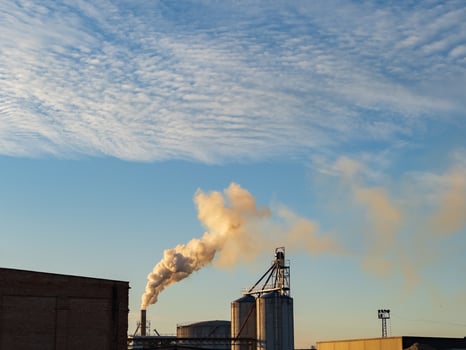 In a sprawling industrial area, smokestacks release plumes of smoke against a backdrop of fluffy clouds. The warm hues of sunset cast a golden light on the structures photo