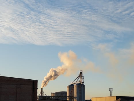As the sun sets, smoke drifts from large industrial silos, painting a dramatic picture against the expansive blue sky. The quiet evening air hints at a bustling production nearby photo