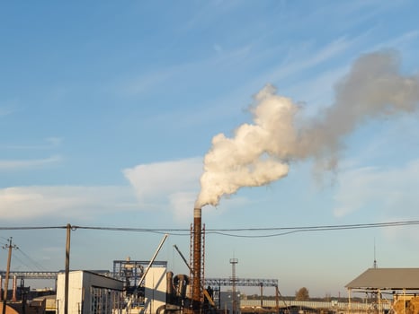 A factory stands tall with smoke rising from its chimney as the sun sets, casting a warm glow over the industrial scene. The sky remains predominantly blue, highlighting the structure photo
