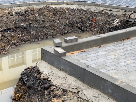An unfinished brick pavement stretches alongside a sidewalk, featuring a trench filled with water. Loose soil and uninstalled pavers indicate ongoing construction activity photo