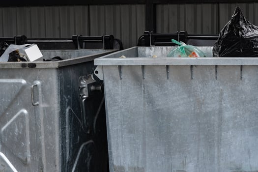 Two metal garbage containers filled with various types of rubbish are situated in a covered public space, collecting waste during daylight hours. The area shows signs of urban disarray photo