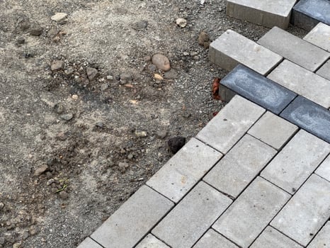 A section of pavement is being constructed, showcasing gray interlocking bricks. Nearby, an unfinished area of gravel and loose stones is visible, indicating progress in development photo