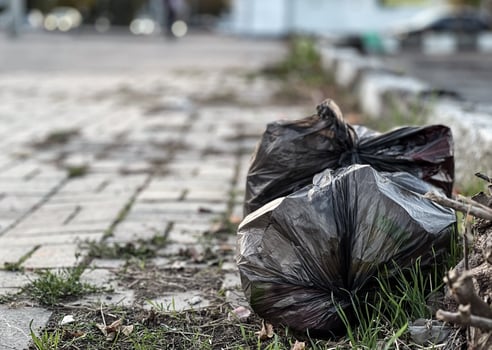 A black plastic bag overflowing with trash rests on the ground next to a brick walkway, highlighting the litter problem in a neglected public space photo