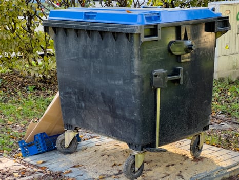 A black trash can with a blue lid rests on a paved city street. The can has four wheels and is made of plastic photo