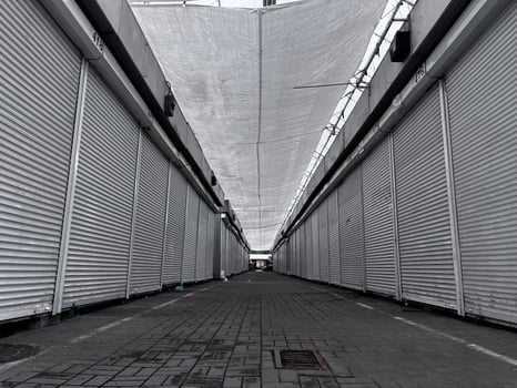 The narrow passageway of a once-busy market stands silent, lined with shuttered stalls. The overhead canopy casts shadows on the vacant ground, hinting at a forgotten vibrancy photo