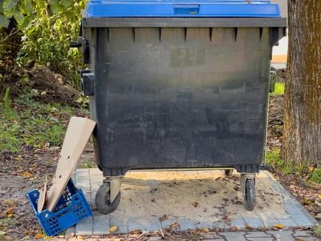 A blue-topped dumpster stands in a serene park, with discarded cardboard and a plastic crate lying nearby, highlighting everyday waste challenges in nature photo