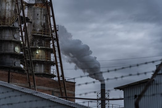 Smoke billows from smokestacks of an industrial facility, surrounded by buildings and fencing, against a backdrop of a moody, overcast evening sky photo