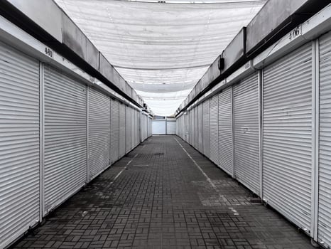 An empty marketplace features rows of closed stalls under a white canopy. The pavement is lined with tiles, and the atmosphere reflects a quiet afternoon in a commercial area photo