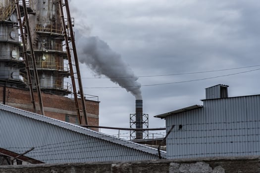 A factory emits thick smoke from its chimney, contrasting against a backdrop of overcast skies and nearby industrial structures, depicting a typical manufacturing environment photo